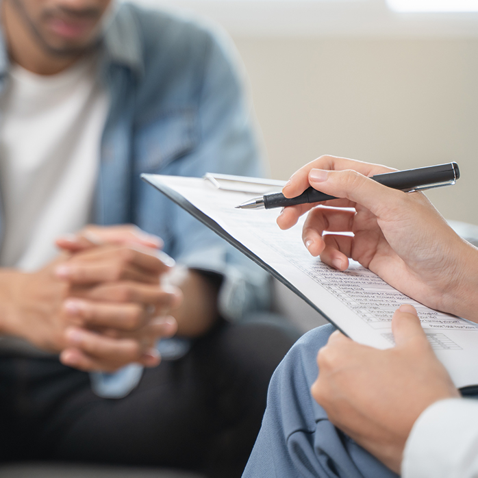 A close-up image of a person holding a pen over a clipboard while another person sits nearby, appearing engaged in a conversation or consultation, possibly in a therapeutic or counseling setting.