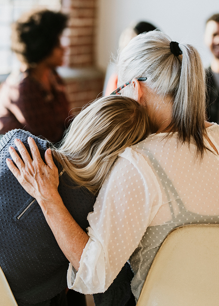 Two women sharing a warm embrace, one resting her head on the other's shoulder, illustrating comfort and connection in a serene setting. 